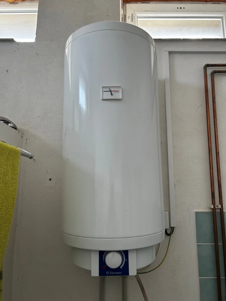 Bílý elektrický bojler na zdi s viditelnými potrubími.