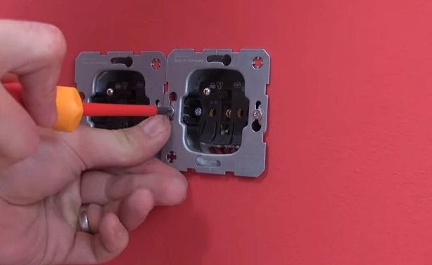 Ruka šroubuje elektrickou zásuvku do zdi pomocí šroubováku.