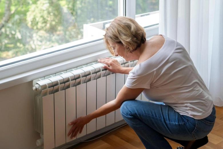 Žena se ohýbá k radiátoru u okna, aby zkontrolovala teplotu.