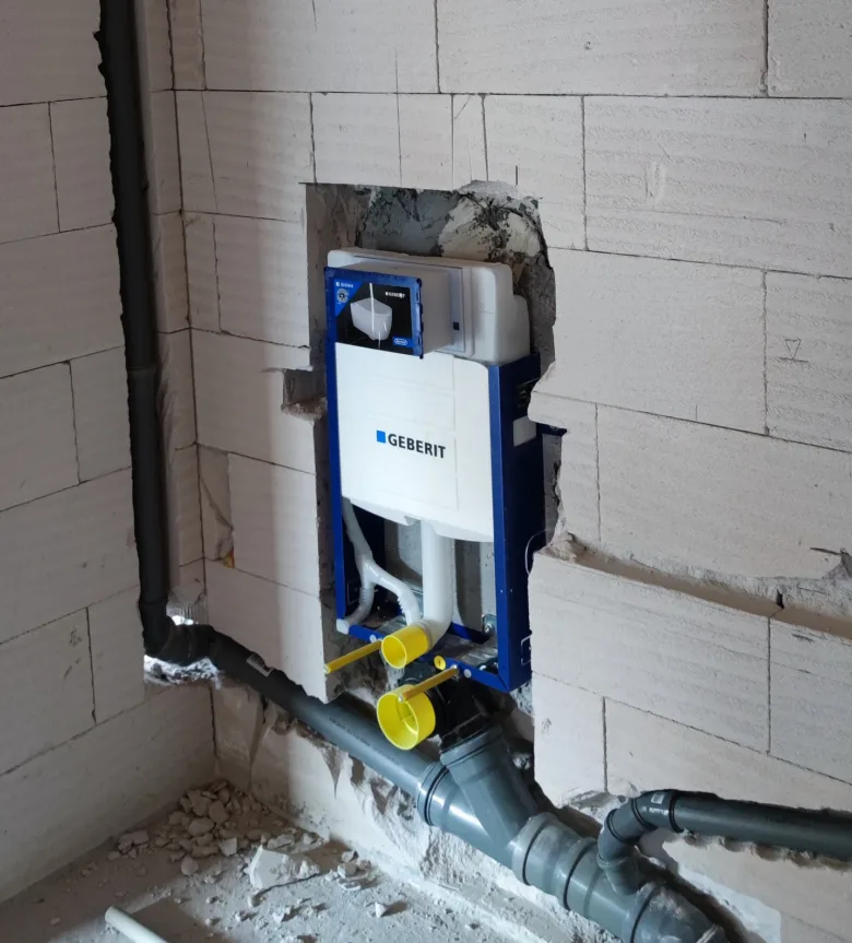 Instalace záchodové nádrže na zeď s viditelnými potrubími a odpady.