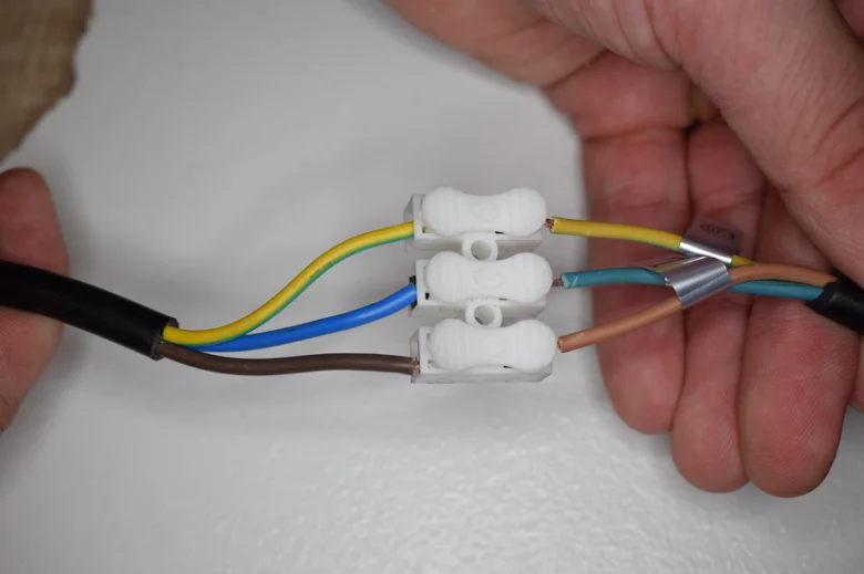 Ruce připojují barevné vodiče k bílému konektoru pro instalaci nástěnné lampy.