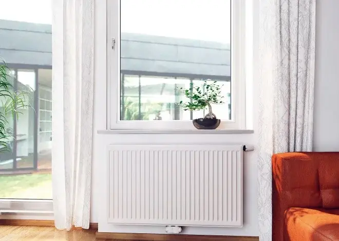 Bílý radiátor pod oknem v moderním obýváku s oranžovou pohovkou.