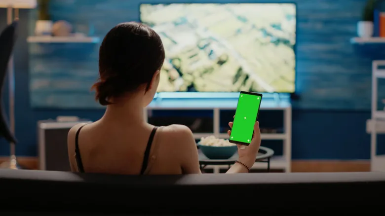 Žena sedí na gauči s telefonem před televizorem v obývacím pokoji.