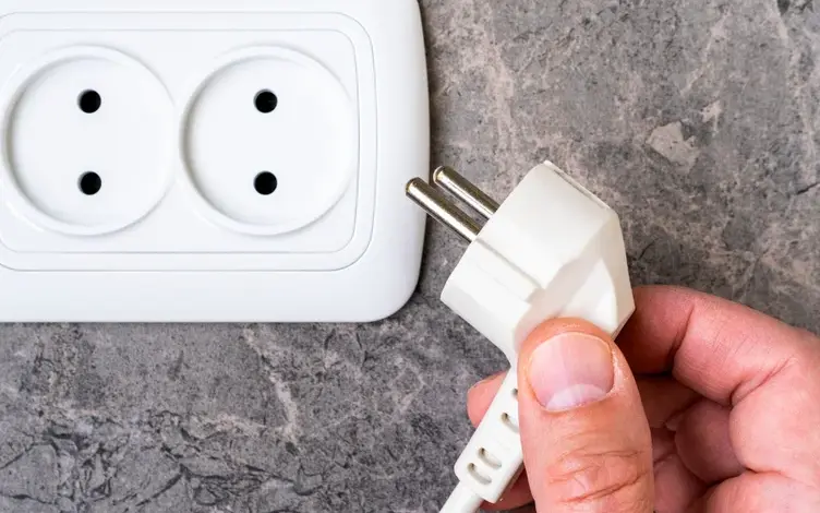 Ruka drží zástrčku u elektrické zásuvky na dlažbě v koupelně.