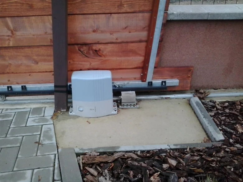 Bílý motor brány u dřevěného plotu s viditelnými mechanickými díly.