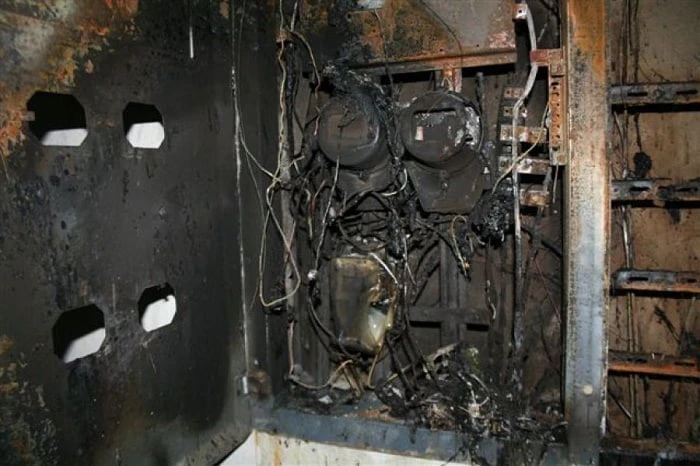 Spálený elektrický rozvaděč s poškozenými vodiči a součástkami uvnitř zdi.