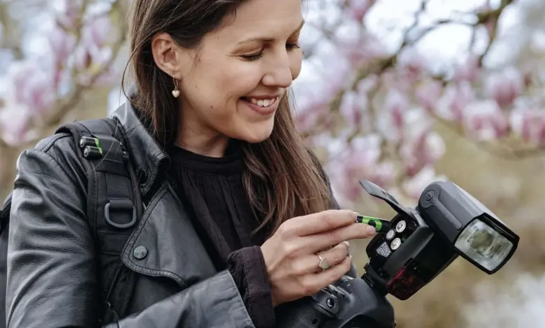 Žena s fotoaparátem vkládá baterii do blesku na pozadí kvetoucích stromů.