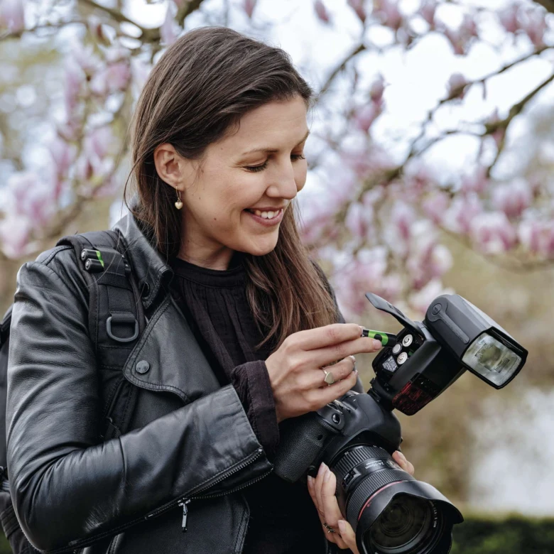Žena vkládá baterie do blesku fotoaparátu na pozadí kvetoucích stromů.