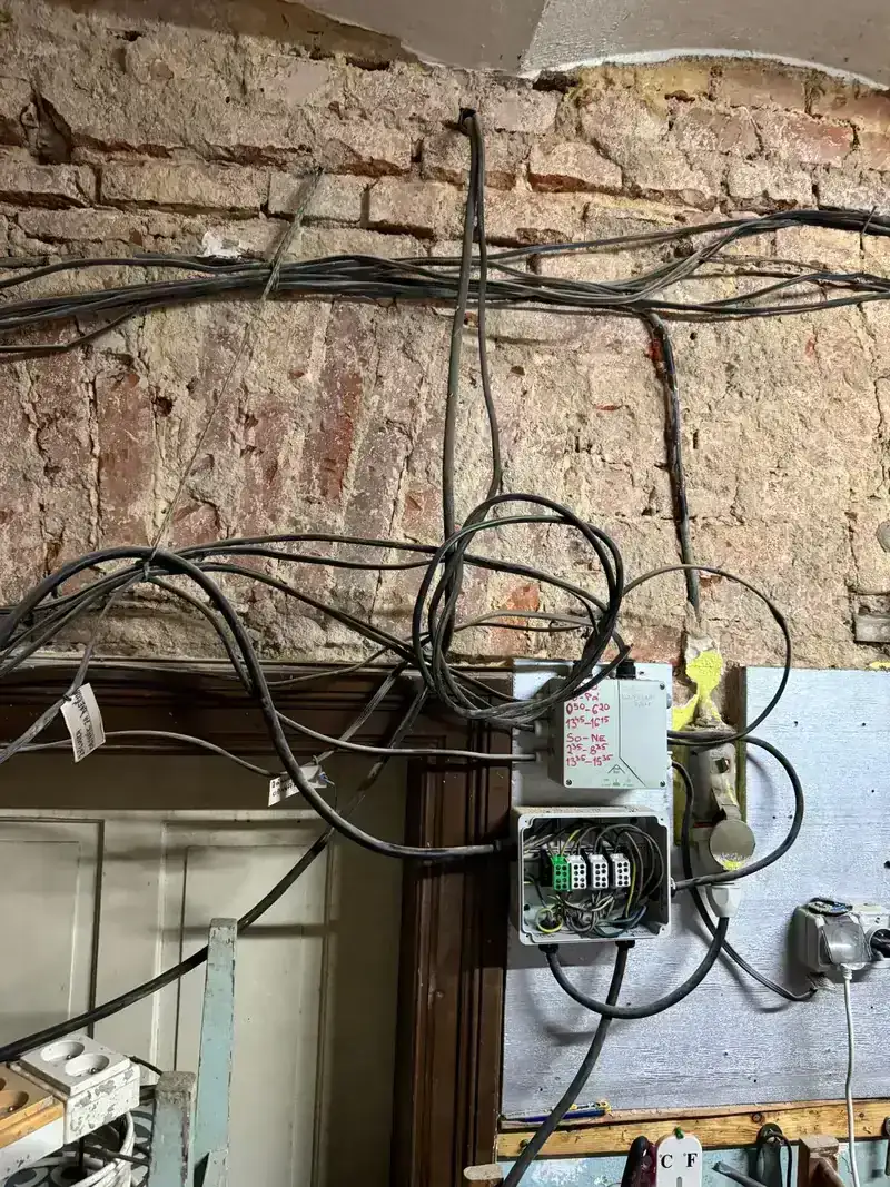 Zamotané elektrické kabely u otevřené zásuvky na cihlové zdi.