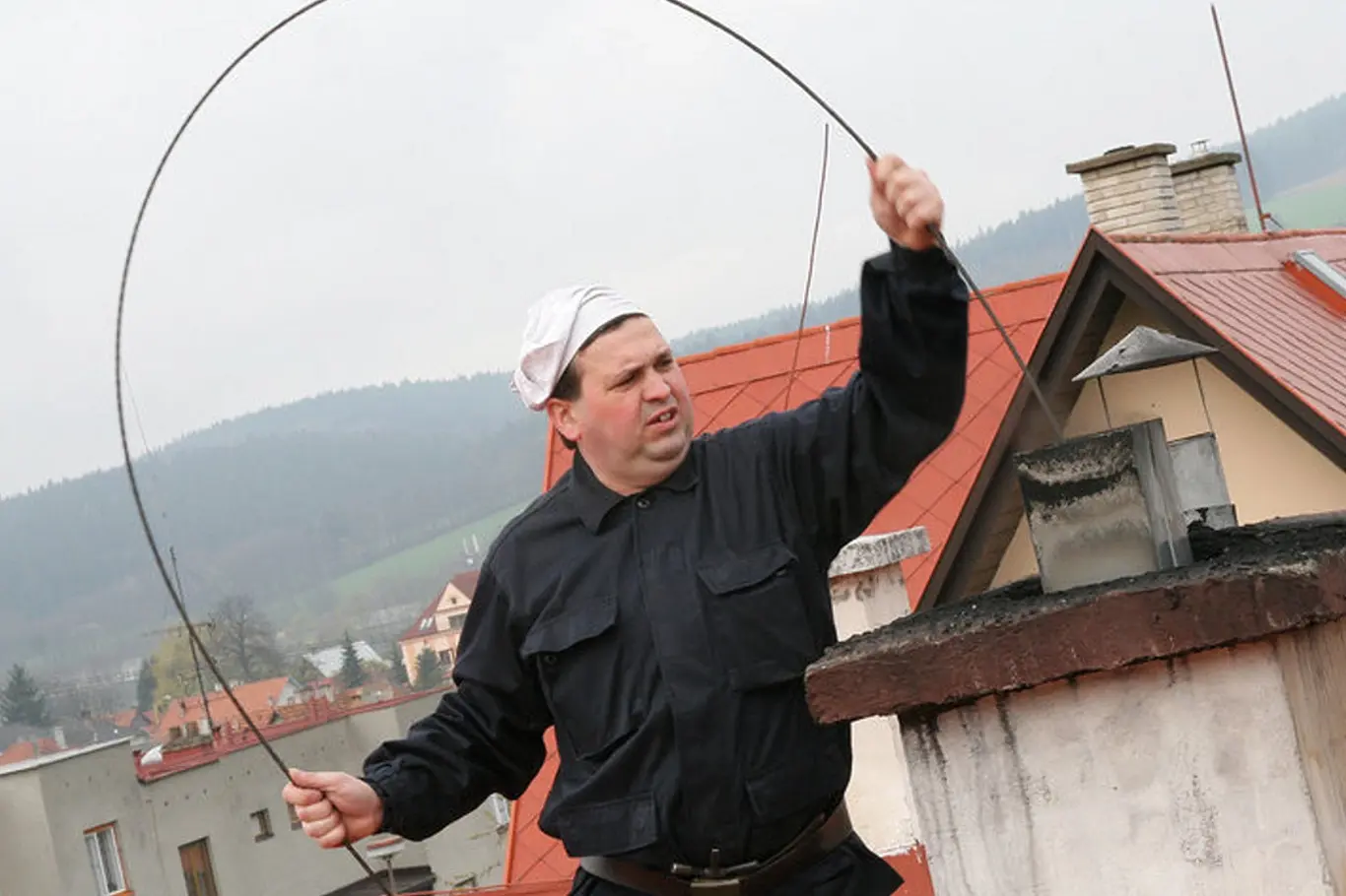 Muž čistí komín dlouhou tyčí na střeše.