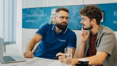 Dva muži diskutují u stolu v kanceláři s klimatizací na pozadí.