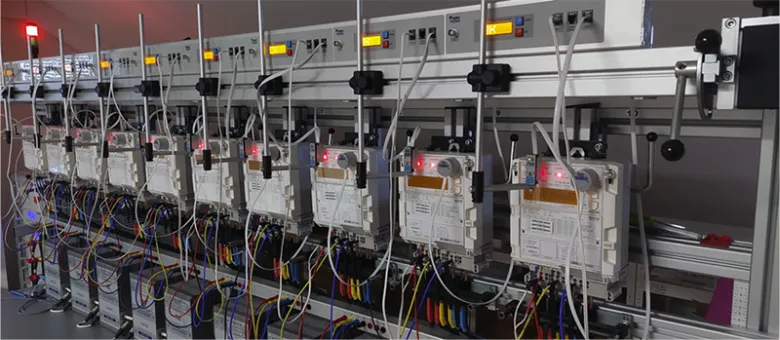 Řada elektroměrů na testovacím stojanu s viditelnými indikátory.