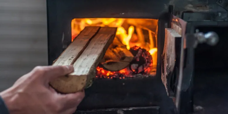 Ruka vkládá dřevo do hořícího kombinovaného kotle na vytápění.