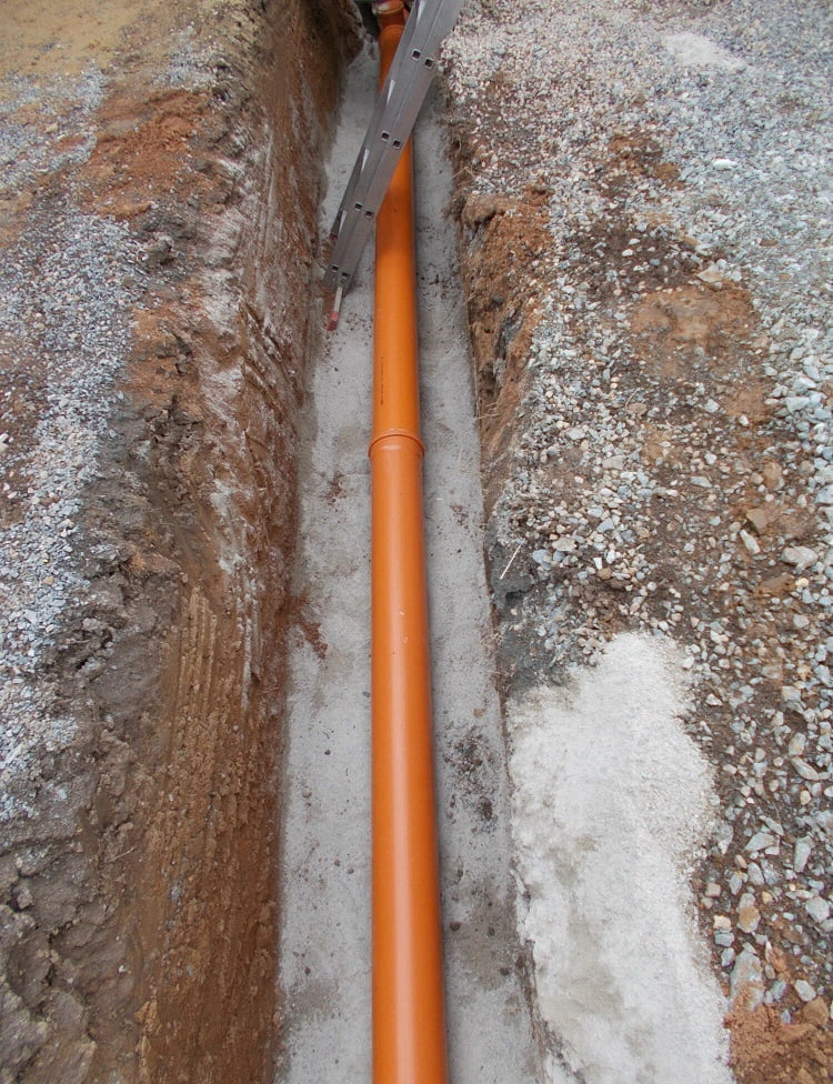 Oranžová kanalizační trubka v příkopu s betonovou podložkou.