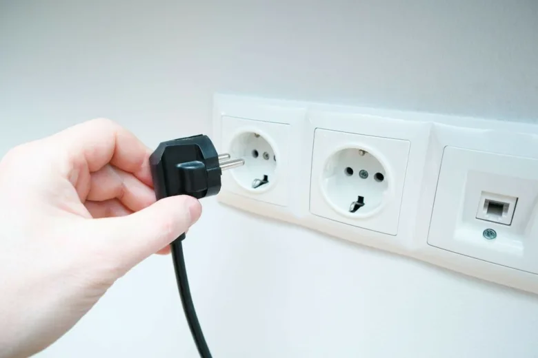 Ruka zapojuje černou zástrčku do elektrické zásuvky na bílé zdi.