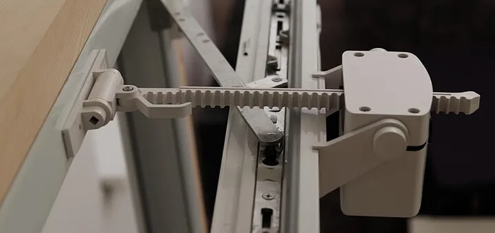 Bílý omezovač otevírání okna připevněný k rámu s viditelným ozubeným mechanismem.