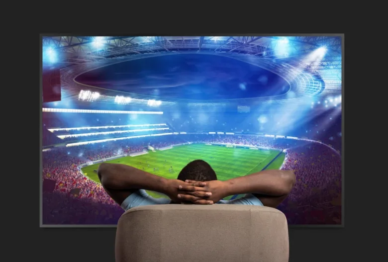 Muž sedí před velkou televizní obrazovkou s fotbalovým stadionem.