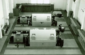 Vnitřní pohled na starou elektrárnu s velkými generátory a ovládacími panely.