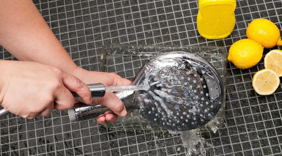Člověk čistí sprchovou hlavu pod proudem vody na dlažbě.