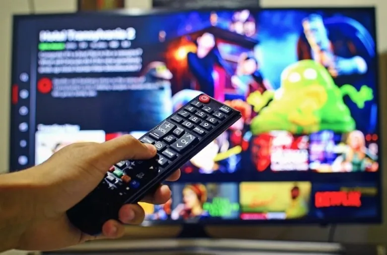 Ruka drží ovladač před televizí s barevným obsahem.