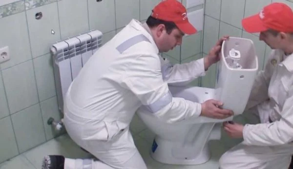 Dva pracovníci montují záchod, připojují nádrž k míse.